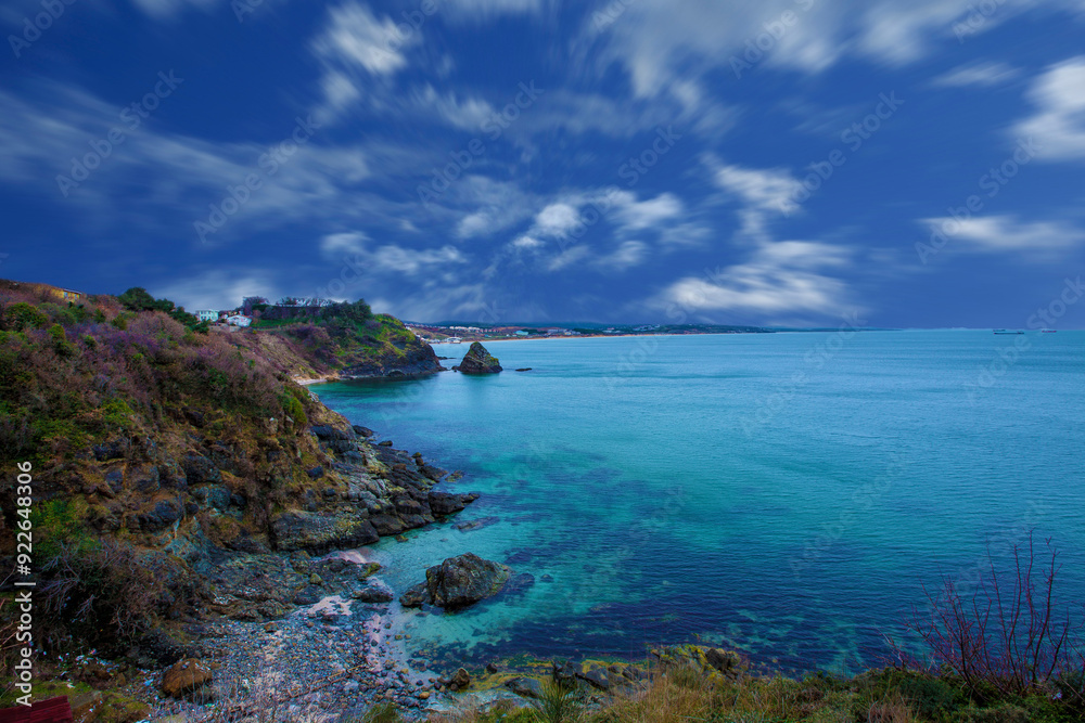 A rocky shore with a body of water and a blue sky with clouds, a body of water surrounded by a lush green hillside A rocky shore with a body of water and a blue sky with clouds, a body of water surrounded by a lush green hillside