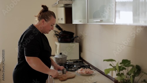 A dark-haired woman in a black T-shirt is preparing homemade chicken wiener schnitzel. She beats the meat with a metal hammer. Cooking in the home kitchen.