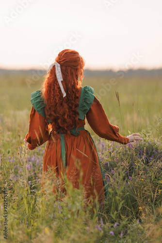 A shepherdess woman with red hair and a red and green dress walks on a field of flowers. Rural scene concept with a romantic peasant woman in a linen dress. Romance novel. photo