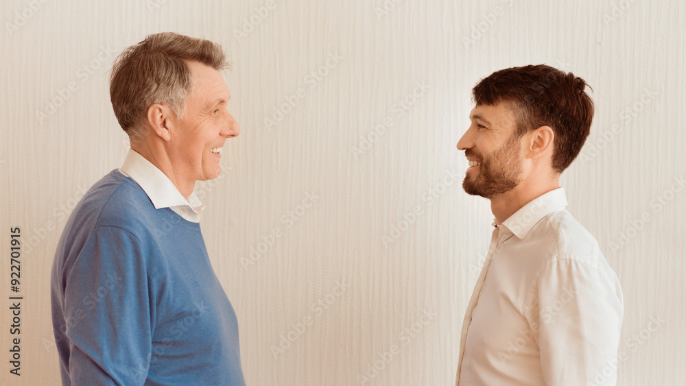Mature Man And His Elderly Dad Smiling Each Other Standing Against White Wall Indoor. Father And Adult Son Relationship. Panorama Mature Man And His Elderly Dad Smiling Each Other Standing Against White Wall Indoor. Father And Adult Son Relationship. Panorama