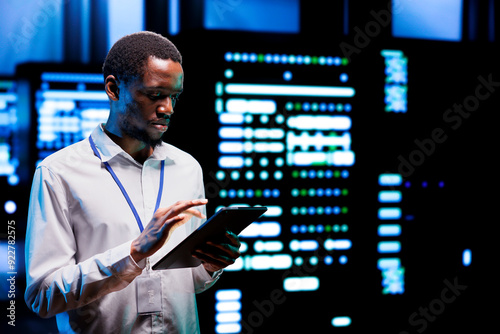 Computer scientist running high tech data center components diagnostic test to determine and patch software issues. African american employee using tablet to mend critical systems checking for defects photo