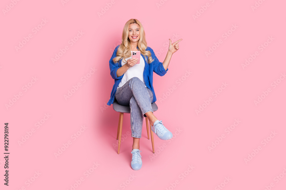 Full size photo of positive woman dressed shirt sit on chair hold smartphone directing empty space isolated on pink color background Full size photo of positive woman dressed shirt sit on chair hold smartphone directing empty space isolated on pink color background