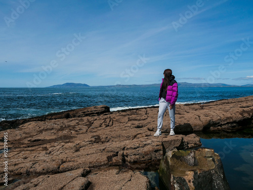 Teenager girl standing on rough stone coast, blue ocean and blue sky in the background. Travel and tourism. Mullaghmore, Ireland. Trip to amazing Irish nature. Girl in purple jacket, in dark glasses. photo