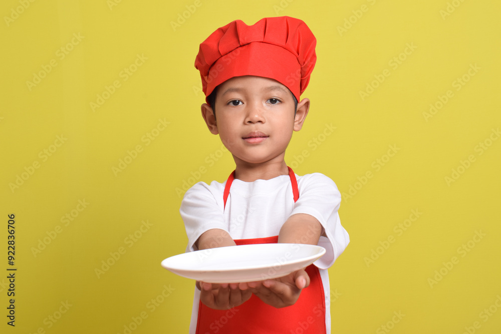 Cute little chef with plate. Asian boy wearing red apron and hat isolated on yellow background. Concept of serving food dishes Cute little chef with plate. Asian boy wearing red apron and hat isolated on yellow background. Concept of serving food dishes