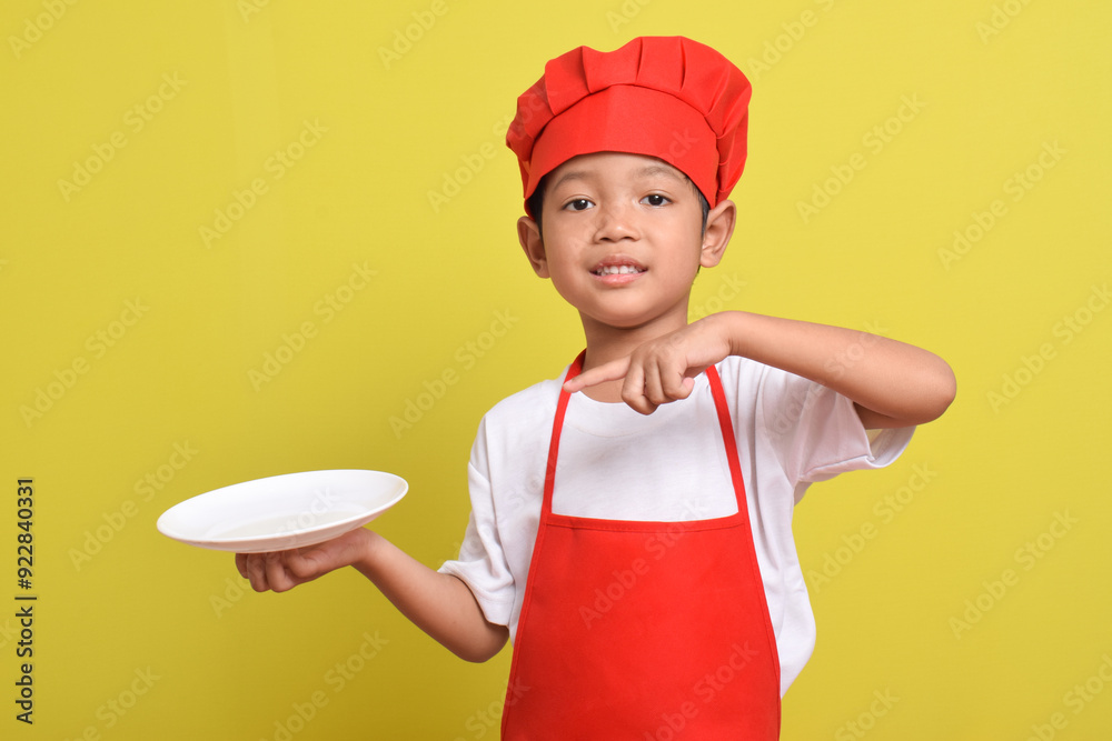 Cute little chef with finger pointing to plate. Asian boy wearing red apron and hat isolated on yellow background. Concept of serving food dishes Cute little chef with finger pointing to plate. Asian boy wearing red apron and hat isolated on yellow background. Concept of serving food dishes
