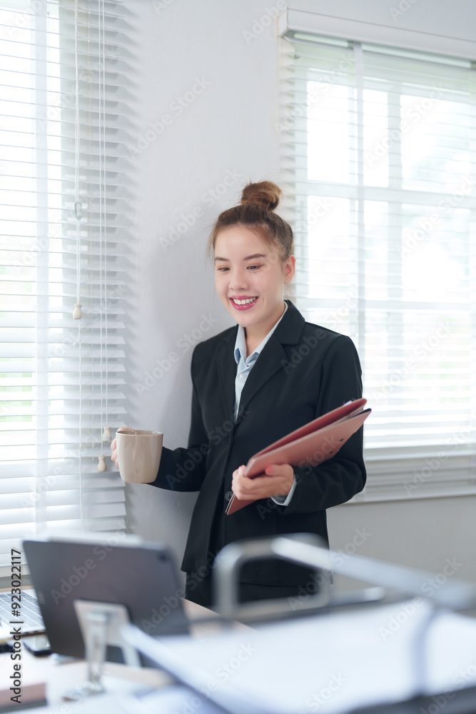 A confident businesswoman in a black suit is standing near a window, holding a coffee cup and a folder, ready to start her day in the office A confident businesswoman in a black suit is standing near a window, holding a coffee cup and a folder, ready to start her day in the office