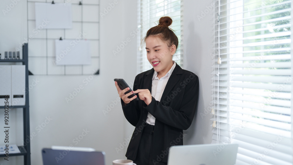 A businesswoman in a black suit is standing by the window, checking her smartphone with a focused expression in a modern office setting A businesswoman in a black suit is standing by the window, checking her smartphone with a focused expression in a modern office setting