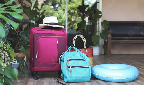travel accessories, pink luggage, woven bag, backpack, headphones, swimming ring  and straw hat on balcony with houseplant.