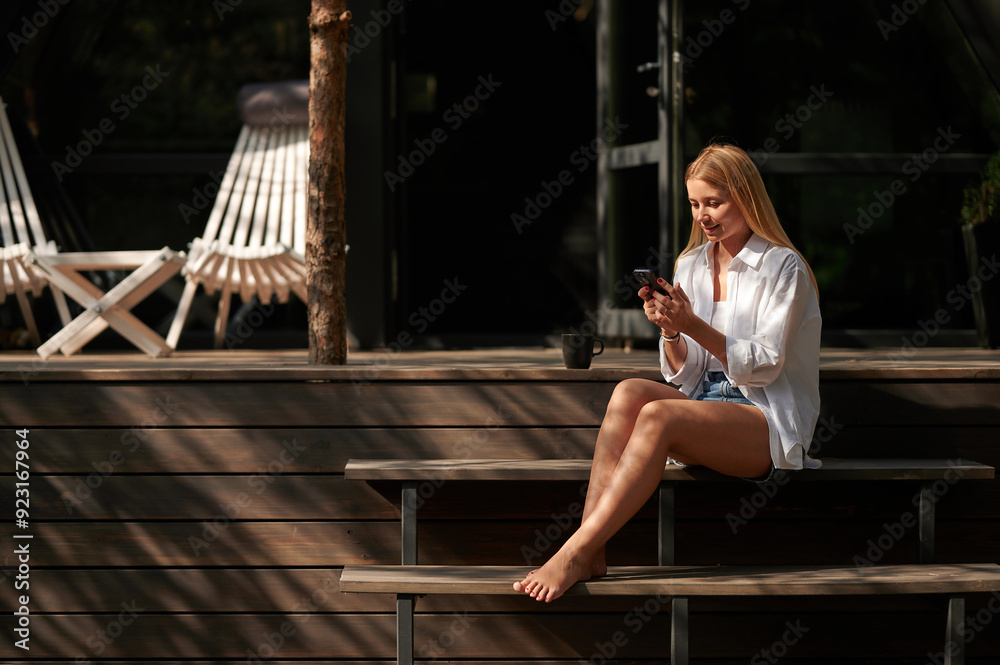 Young pretty girl in the rays of the summer setting sun reading the news with a smartphone sitting on the porch of an A-frame house. Young pretty girl in the rays of the summer setting sun reading the news with a smartphone sitting on the porch of an A-frame house.