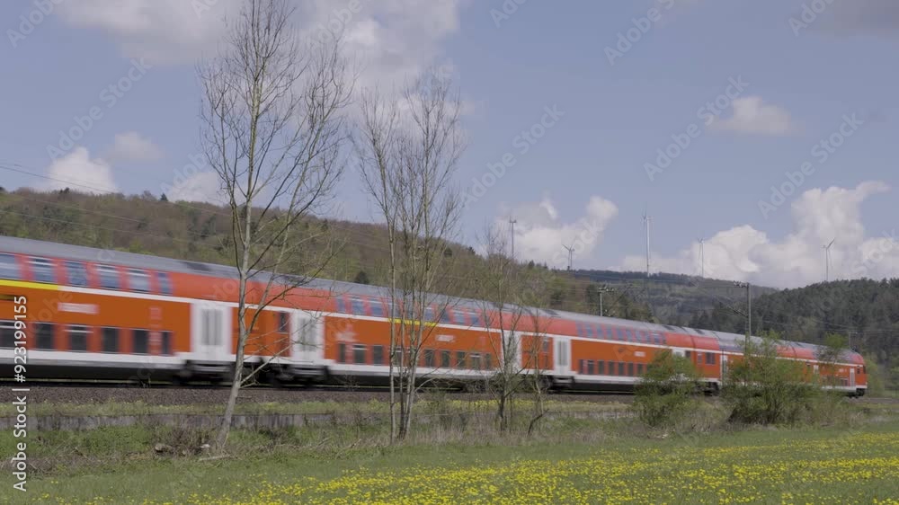 An orange train speeds through a scenic countryside with wind turbines and trees in the background An orange train speeds through a scenic countryside with wind turbines and trees in the background