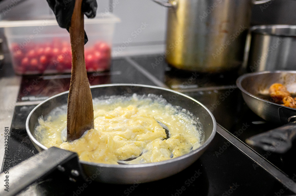 Close-up of scrambled eggs being cooked in a frying pan with a wooden spatula, on a stove with other cookware in the background. Perfect for breakfast, cooking, and kitchen-related themes.