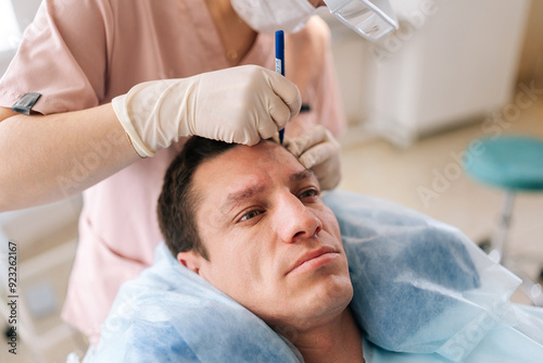 Closeup of unrecognizable female nurse drawing using pencil new hairline on head of handsome patient male before hair transplantation surgery in clinic. Concept of professional baldness treatment.