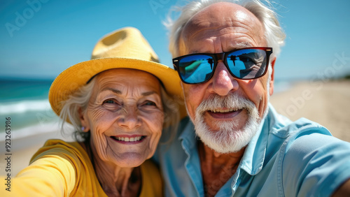 Smiling elderly couple enjoying a sunny day at the beach, capturing the moment with a cheerful selfie, both dressed in casual clothing and sunglasses, with the ocean in the background.