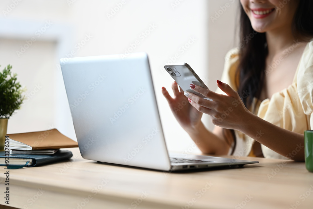Smiling young woman at working desk with her laptop and using mobile phone Smiling young woman at working desk with her laptop and using mobile phone