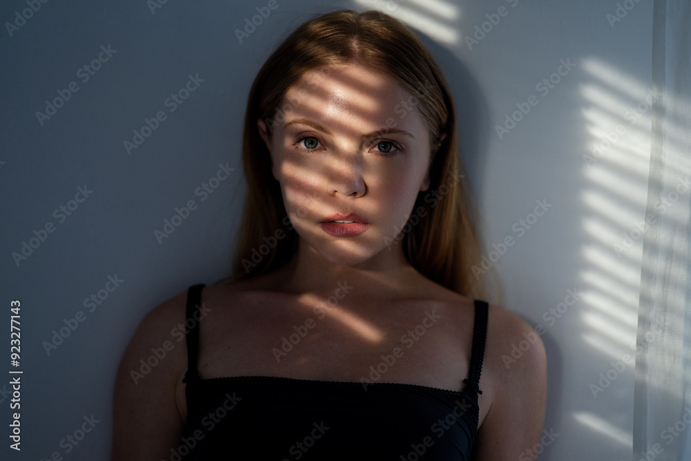 Fototapeta premium Young Woman looking through window blinds. Portrait of a woman in shadows on face from window blinds. Girl pensive look at window in living room. Portrait female model with shadow from the blinds.