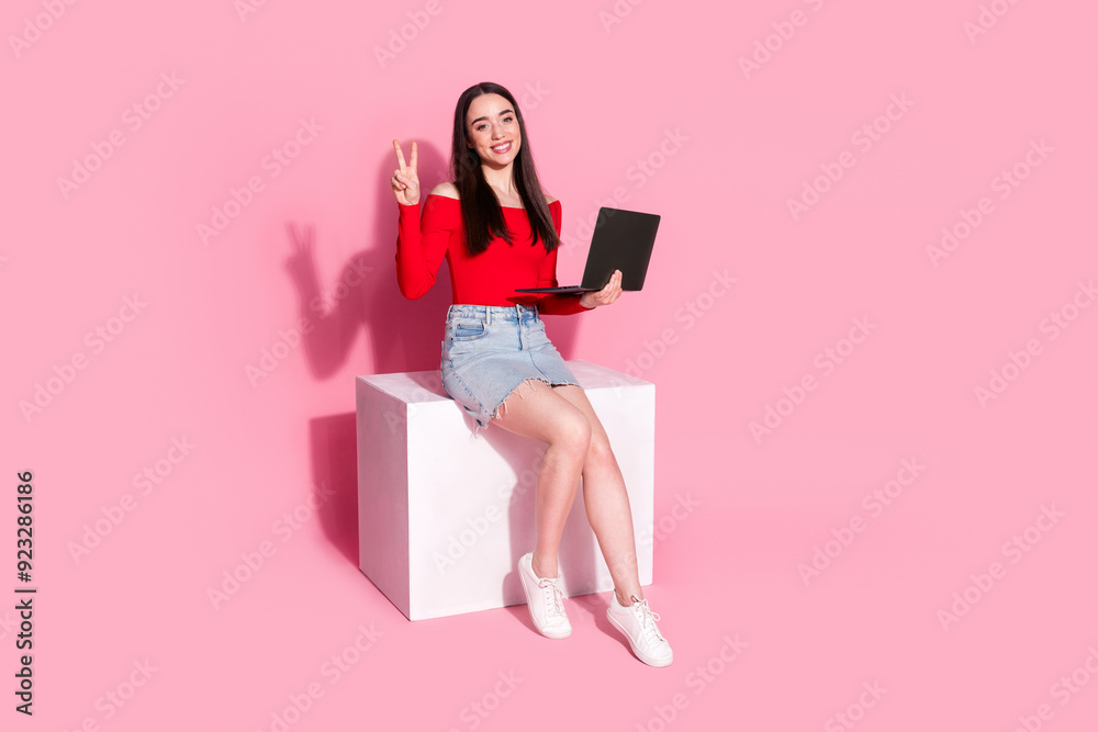 Full size photo of gorgeous young woman sit podium laptop v-sign isolated on pink color background Full size photo of gorgeous young woman sit podium laptop v-sign isolated on pink color background