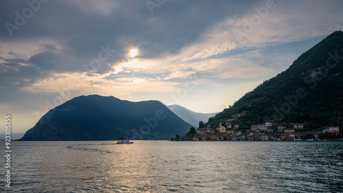 A tranquil scene of a village nestled on the shores of Lago d'Iseo, with mountains in the background and a soft evening light illuminating the landscape.