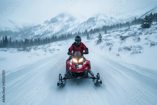 A person riding a red snowmobile at high speed on a snow-covered mountain trail with pine trees and snowy peaks in the background, showcasing winter adventure. photo