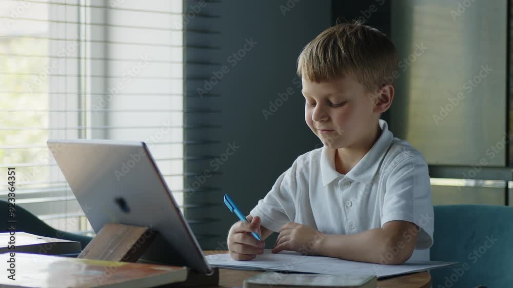 Portrait. A child is studying in an online school using a tablet while sitting at home in the kitchen at the table and writes with a fountain pen in a notebook. Portrait. A child is studying in an online school using a tablet while sitting at home in the kitchen at the table and writes with a fountain pen in a notebook.