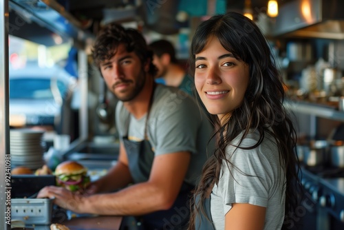 Smiling woman with long black hair standing next to a man preparing a burger in a food truck.
