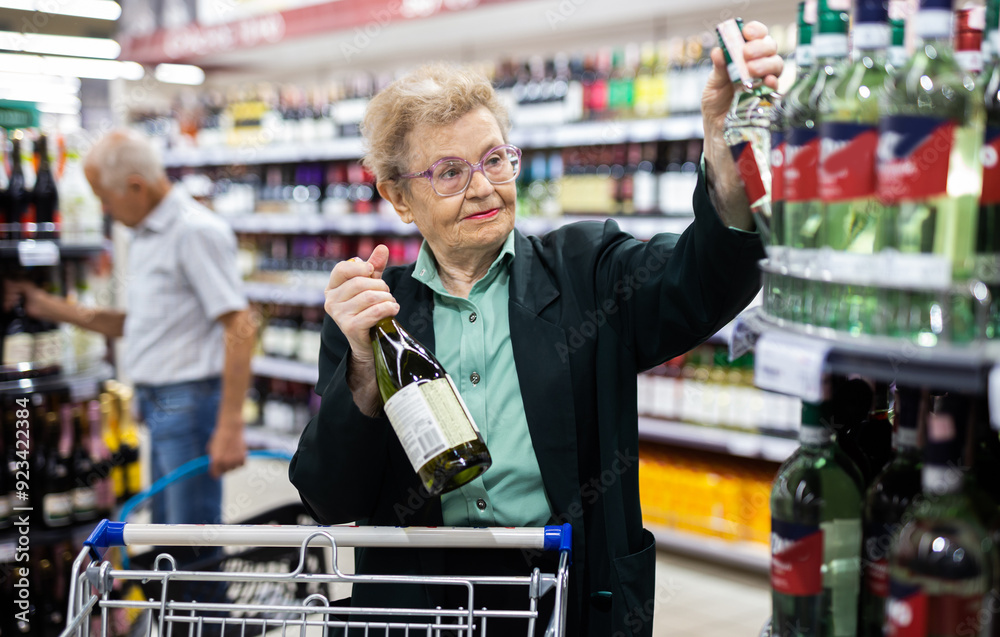 Φωτογραφία Mature woman with glasses chooses bottle of wine in alcohol section of supermark