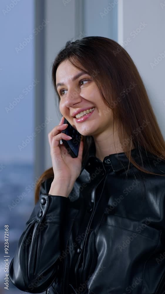 Smiling woman stands at the window speaking on the phone. Brunette businesswoman having business conversation. Smiling woman stands at the window speaking on the phone. Brunette businesswoman having business conversation.