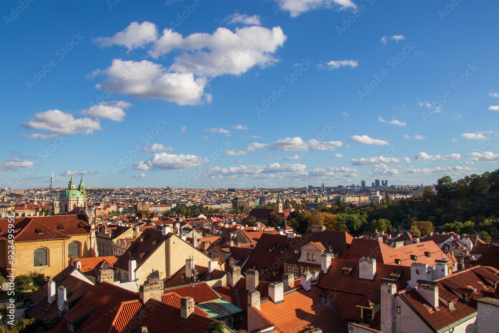Panoramic View of Prague with Red Roofs and Blue Sky. Panoramic view of a European city with red-tiled roofs, church spires, lush greenery, and a bright blue sky with clouds. Panoramic View of Prague with Red Roofs and Blue Sky. Panoramic view of a European city with red-tiled roofs, church spires, lush greenery, and a bright blue sky with clouds.