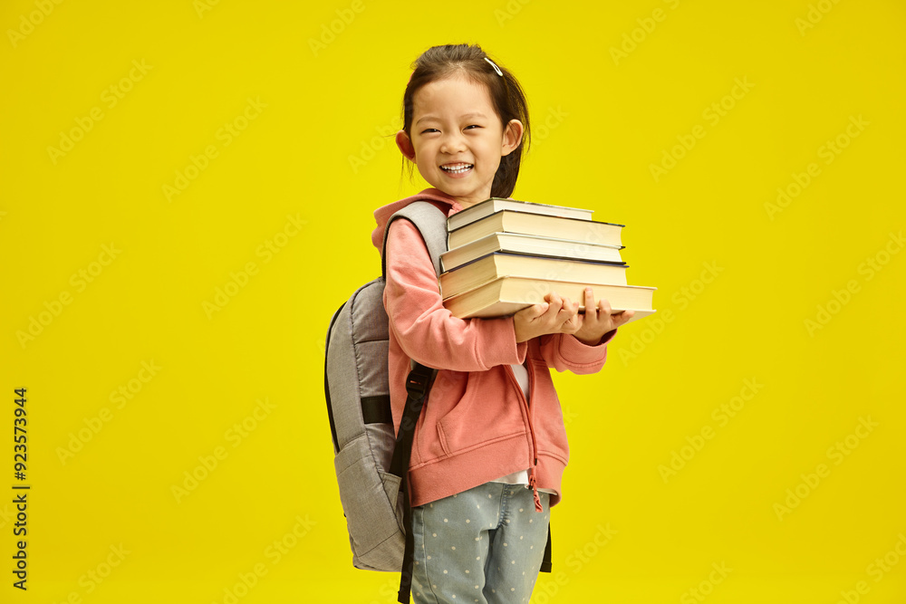 Happy chinese girl preparing going to the first grade, holds many different books in her hands and having backpack on shoulders posing on yellow isolated. Children education in preschool concept. Happy chinese girl preparing going to the first grade, holds many different books in her hands and having backpack on shoulders posing on yellow isolated. Children education in preschool concept.