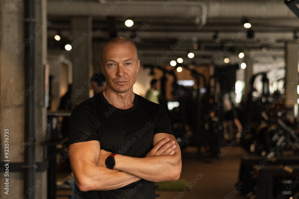 Handsome adult personal trainer or fitness coach standing wearing black t-shirt with cross hand at gym. Motivation for regular workout, body building, health and energy. Crafting my workout routine.