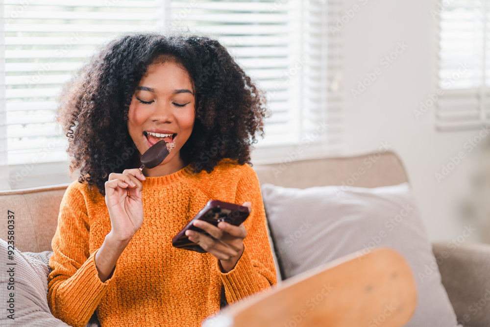 African American woman eating ice cream bar while browsing on smartphone at home. African American woman eating ice cream bar while browsing on smartphone at home.