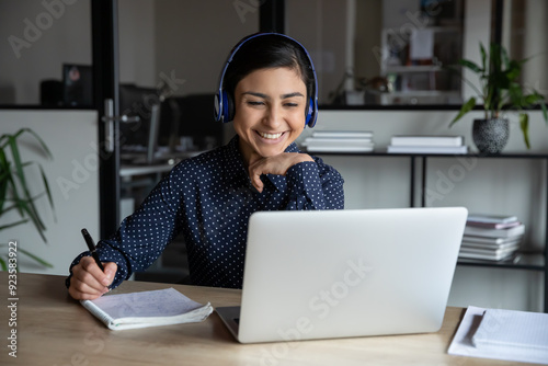 Smiling millennial Indian female employee in wireless earphones make notes watching webinar on laptop, happy young ethnic woman worker in headset talk on video call brainstorm with colleagues online