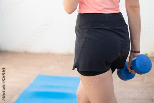 Young woman holding a blue dumbbell during an outdoor workout, fitness training on a yoga mat, wearing athletic shorts and tank top, strength exercise, healthy lifestyle, workout from home.