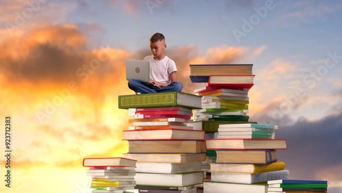 A young boy sits on a stack of books and types on a laptop keyboard