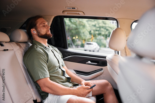 Relaxed man enjoying music in back seat of a car with earphones on, surrounded by a comfortable interior