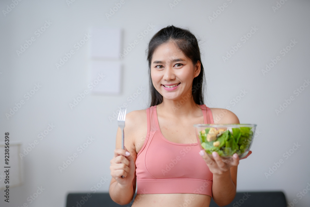 Young asian woman is smiling while holding a bowl of salad and a fork after a workout at home Young asian woman is smiling while holding a bowl of salad and a fork after a workout at home