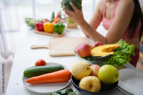 Young woman is holding broccoli in her hands, surrounded by a variety of fresh fruits and vegetables on a white kitchen table