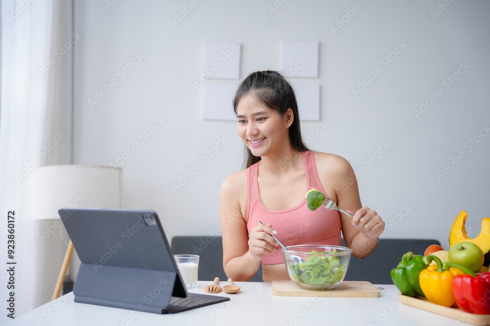 Smiling young asian woman in sportswear is eating a healthy salad and watching an online fitness course on a tablet at home Smiling young asian woman in sportswear is eating a healthy salad and watching an online fitness course on a tablet at home