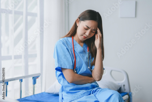 Young doctor in blue scrubs is sitting on a hospital bed with her hand on her head, experiencing a headache and stress