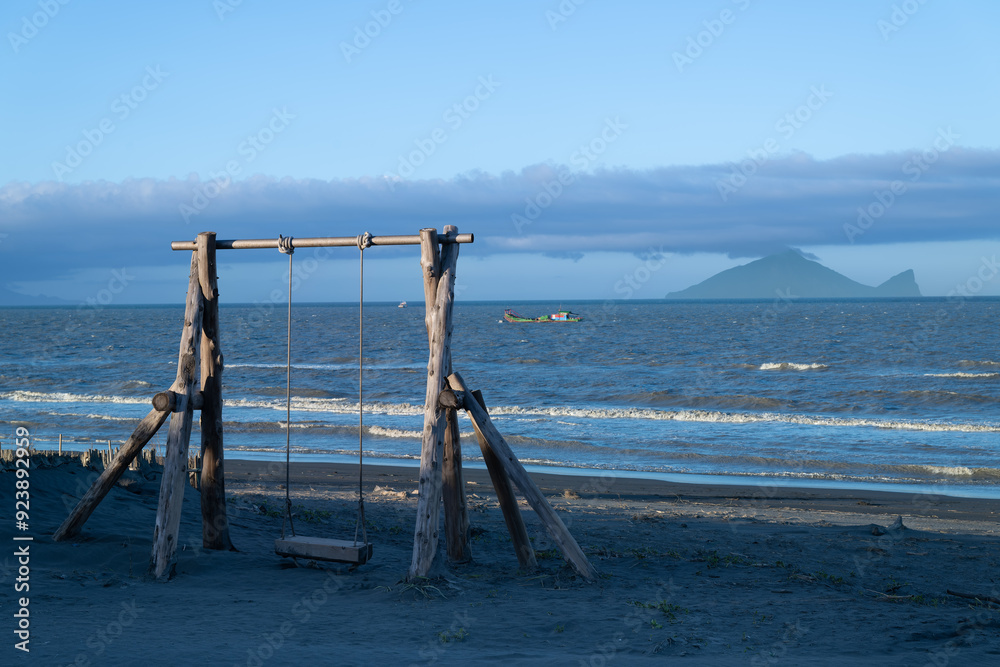 A solitary wooden swing set beckons on a tranquil beach. In the distance, the Turtle Island is partially veiled by clouds, creating a serene coastal landscape. Zhuangwei, Yilan County, Taiwan. A solitary wooden swing set beckons on a tranquil beach. In the distance, the Turtle Island is partially veiled by clouds, creating a serene coastal landscape. Zhuangwei, Yilan County, Taiwan.