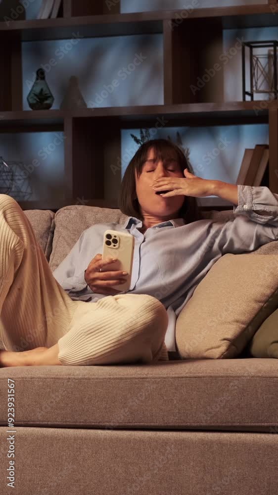 Sleepy Caucasian middle aged woman using mobile phone for messaging in online social media and yawning, lying on couch at home. Vertical video. Sleepy Caucasian middle aged woman using mobile phone for messaging in online social media and yawning, lying on couch at home. Vertical video.