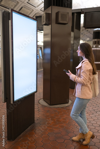 Metro station, a young woman looking at a wall map for directions, holding a cellphone with copy space.