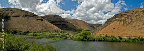 A panoramic view of a portion of the Yakima River Canyon with a footbridge across the river and the brown hills in the background with clouds and blue sky in summer_07072009_0002.