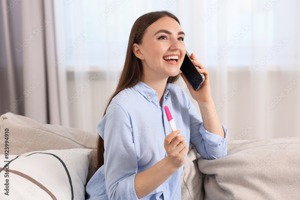 Happy woman with pregnancy test talking on smartphone indoors