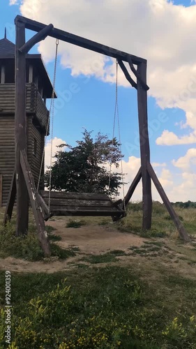 Empty vintage wooden swing in the shape of a bench sways gently in the wind near the Watchtower in Opishnya, Poltava region, Ukraine, amidst lush summer greenery. Vertical 4K video, 28 seconds photo