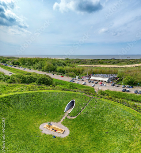 An aerial photo of a large green artificial crater created in the dunes of Kijkduin in The Hague with a tunnel opening in a coastal landscape. 