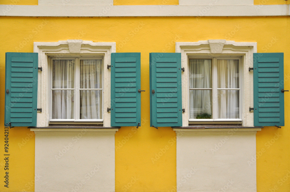 Fototapeta premium Bright yellow wall with two windows featuring teal shutters