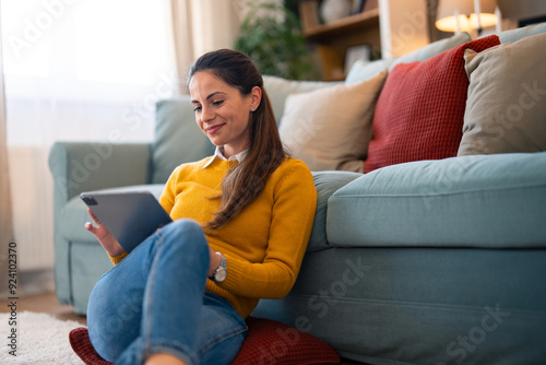 A cheerful woman in casual attire enjoys browsing on her digital tablet while comfortably seated on the floor against a couch in a well-lit living room.