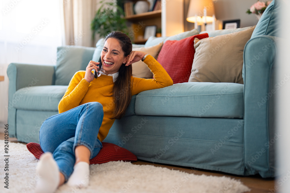 A cheerful caucasian woman enjoys a conversation on her mobile phone while relaxing on the floor near a sofa, her casual attire adding a cozy touch to the bright living room setting. A cheerful caucasian woman enjoys a conversation on her mobile phone while relaxing on the floor near a sofa, her casual attire adding a cozy touch to the bright living room setting.