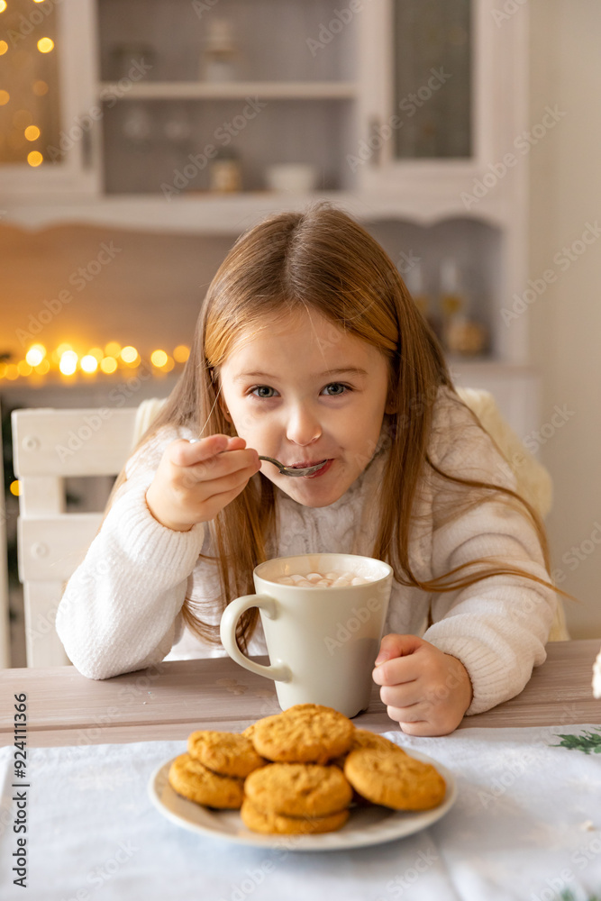 A little girl drinks cocoa with marshmallows in the kitchen at home on Christmas day. Cozy house. Family day off. The concept of the new year. A little girl drinks cocoa with marshmallows in the kitchen at home on Christmas day. Cozy house. Family day off. The concept of the new year.