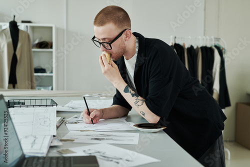 Young busy male fashion designer with pencil biting sandwich and drawing sketch of attire for new seasonal collection by workplace photo